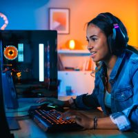 Pretty and Excited Black Gamer Girl in Headphones is Playing First-Person Shooter Online Video Game on Her Computer. Room and PC have Colorful Neon Led Lights. Cozy Evening at Home.