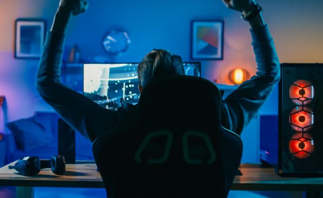 Back Shot of a Gamer Playing and Winning in First-Person Shooter Online Video Game on His Powerful Personal Computer. Room and PC have Colorful Neon Led Lights. Cozy Evening at Home.