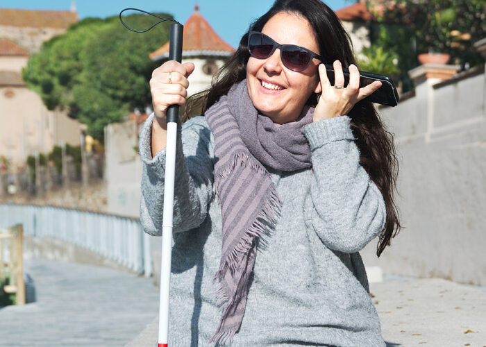 Bild, eine Frau mit Sehbehinderung sitzt im Freien mit ihrem Stock in der rechten Hand und ihrem Mobiltelefon in der linken Hand