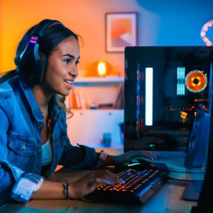 Pretty and Excited Black Gamer Girl in Headphones is Playing First-Person Shooter Online Video Game on Her Computer. Room and PC have Colorful Neon Led Lights. Cozy Evening at Home.