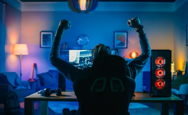 Back Shot of a Gamer Playing and Winning in First-Person Shooter Online Video Game on His Powerful Personal Computer. Room and PC have Colorful Neon Led Lights. Cozy Evening at Home.