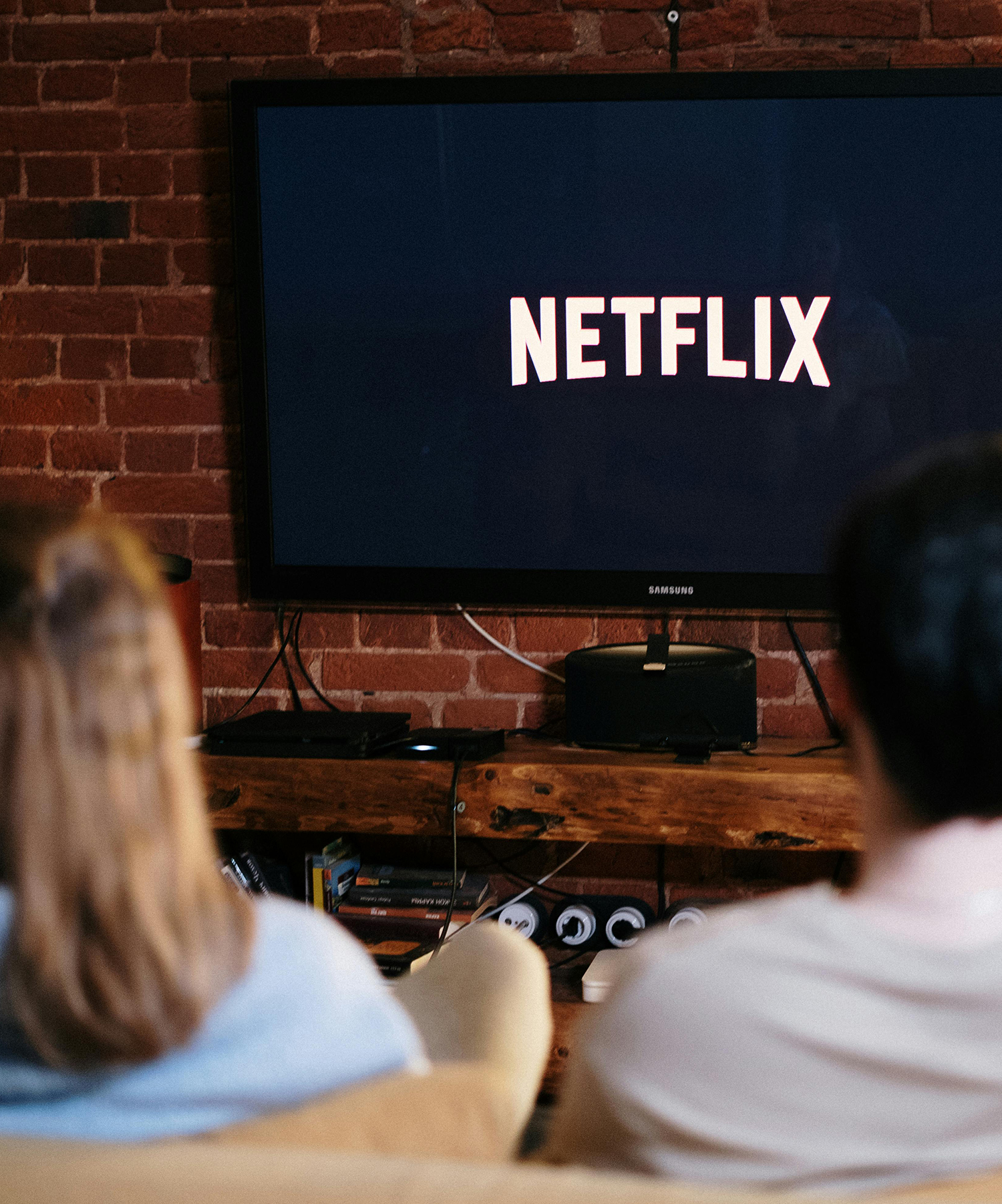 Two people sitting in front of a TV on a couch. The TV has the Netflix title screen on it.